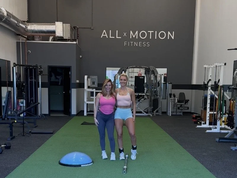 Allie and Monica inside All x Motion Fitness, with the logo on the wall behind them